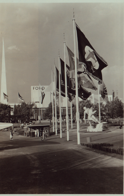 Further away view of the statue, with transportation-related flags in the foreground and the ford building in the background