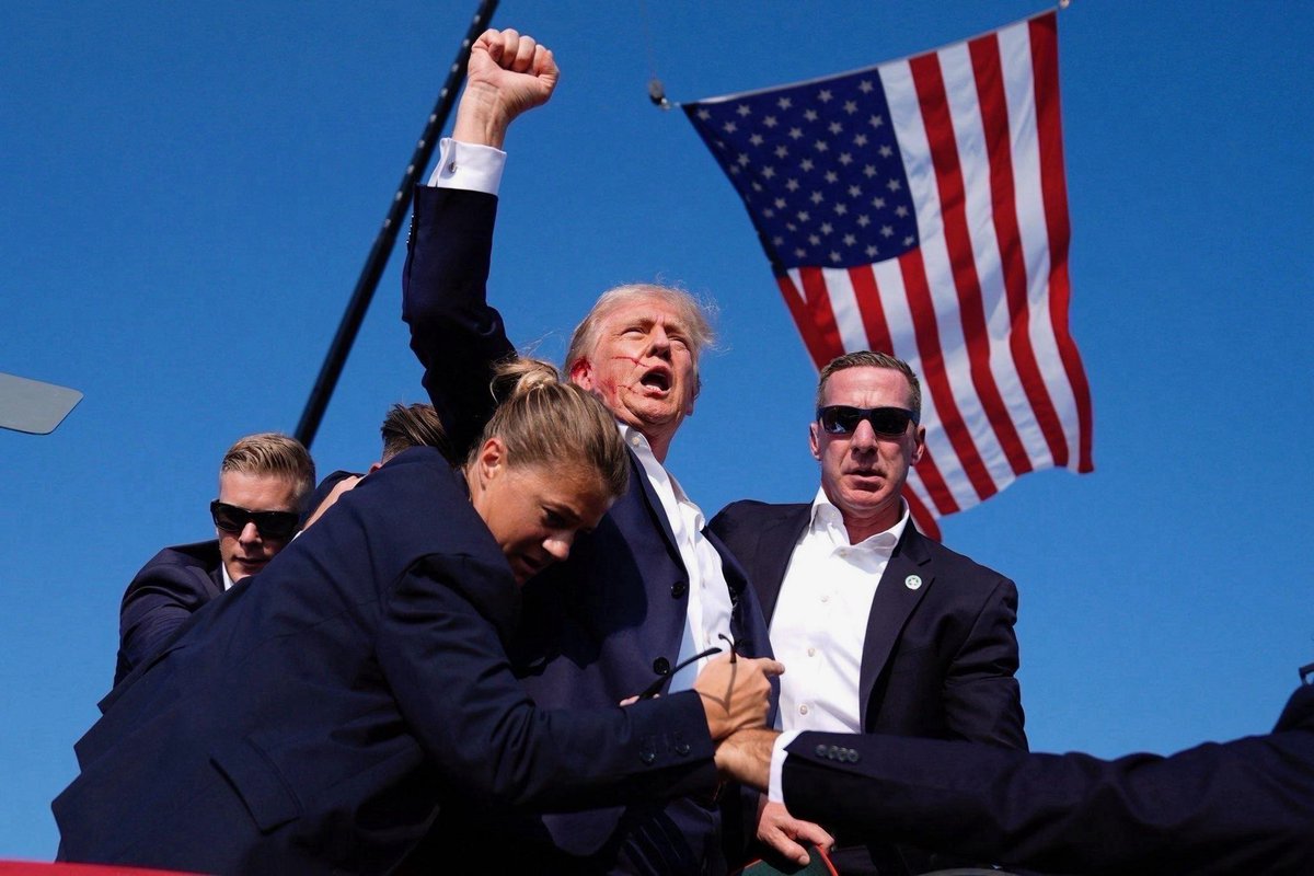 Trump surrounded by bodyguards slightly bloodied near his right ear and pumping a fist in front of a US flag