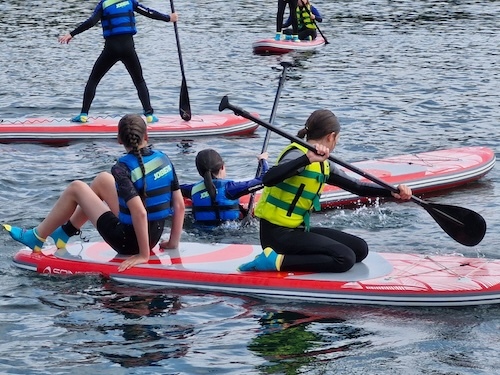 Young carers in wetsuits, paddling on paddle boards on a lake.