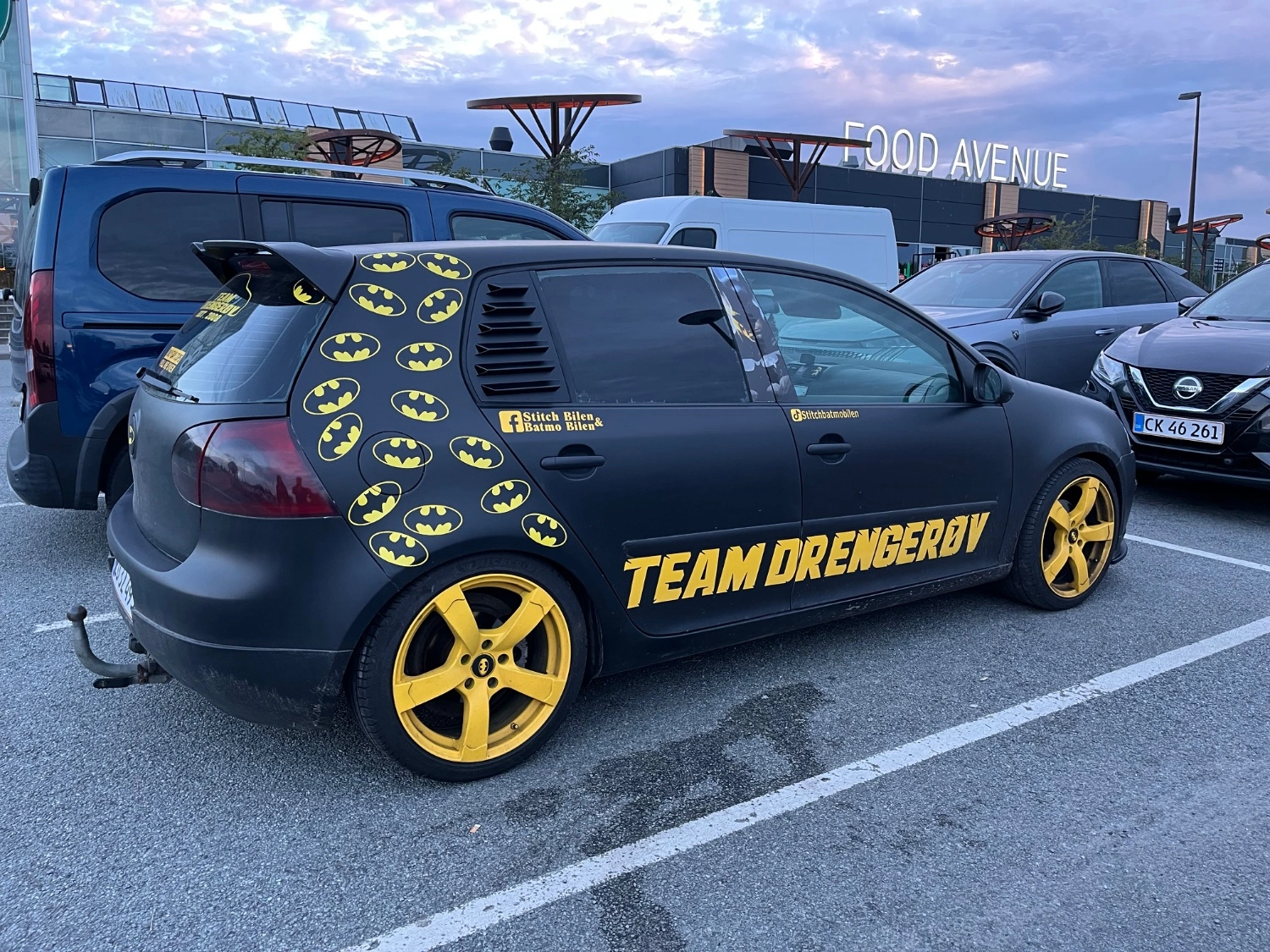Bilen på billedet er en sort personbil med iøjnefaldende gule fælge og flere Batman-logoer på siden. Den bærer teksten "TEAM DRENGERØV" i store gule bogstaver, hvilket giver den et unikt og tematisk udtryk. Bilen er tydeligt dekoreret med et superhelte-inspireret design og skiller sig markant ud i bybilledet.